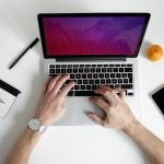 Overhead view of two hands typing on a laptop with a purple screen, beside a notebook, a pen, a phone and an orange on a white desk.