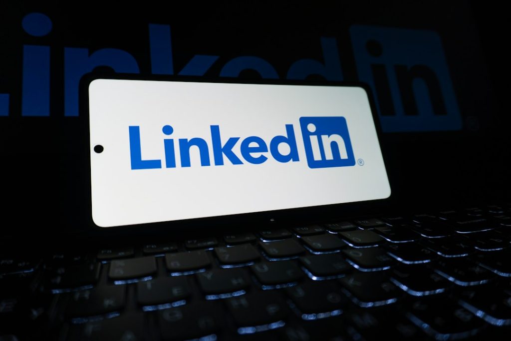 A smartphone standing on a backlit laptop keyboard in the dark, its white screen showing the blue LinkedIn logo.