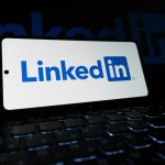 A smartphone standing on a backlit laptop keyboard in the dark, its white screen showing the blue LinkedIn logo.