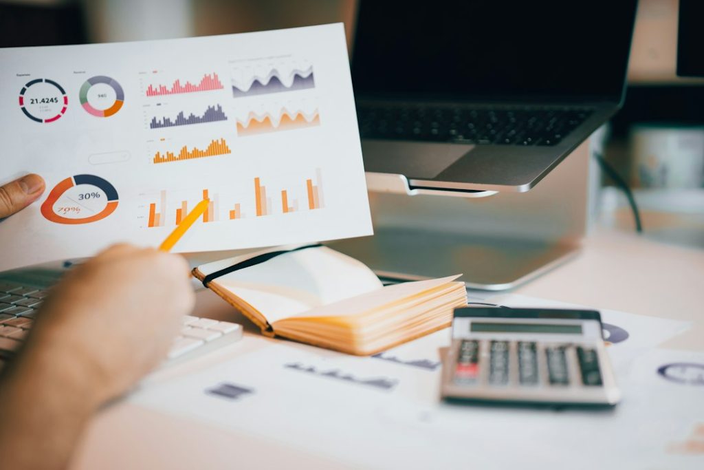 A hand holds a printed page of colorful bar charts and donut charts over a desk with an open notebook, a calculator and a laptop on a stand.