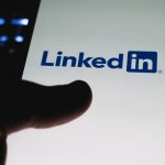 A dark silhouette of a thumb hovering over a phone screen that shows the LinkedIn logo on a white background.