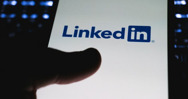 A dark silhouette of a thumb hovering over a phone screen that shows the LinkedIn logo on a white background.